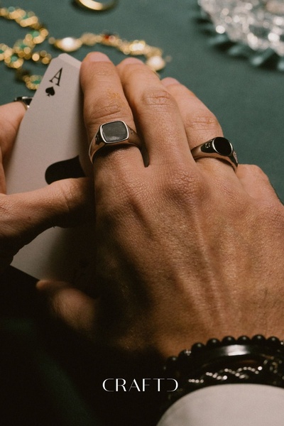 A close-up photograph of a hand wearing two signet rings while holding a card, evoking a sense of luxury, risk, and classic style.