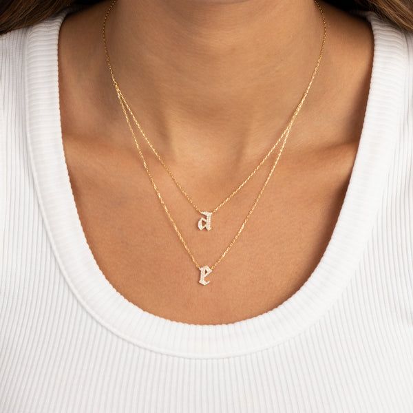 A close-up studio shot of a model wearing two layered gold initial necklaces. The focus is on the jewelry against a clean, neutral background.
