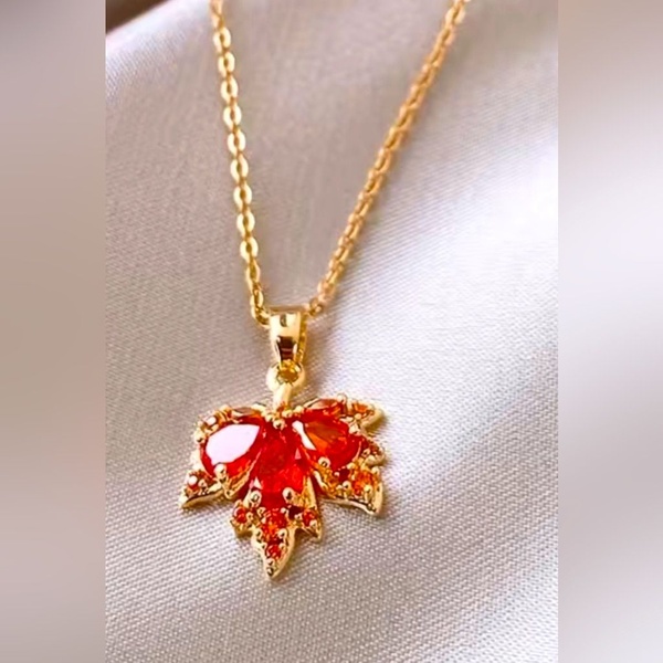 A close-up product shot of a gold-toned maple leaf pendant with vibrant orange gemstones resting on a soft-textured fabric.