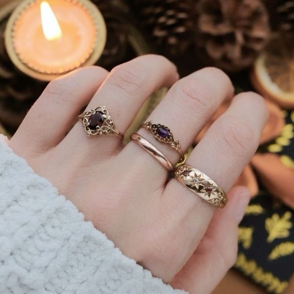 A close-up, warm-toned hand wearing multiple rings, set against a softly lit background with a candle and pine cones for a seasonal, intimate feel.