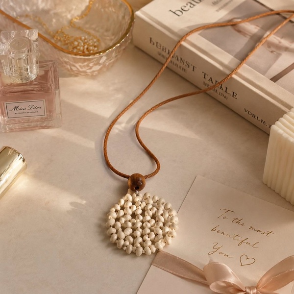 A high-aesthetic, soft-lit product shot of a minimalist pendant necklace styled on a marble surface with perfume and a gift card.