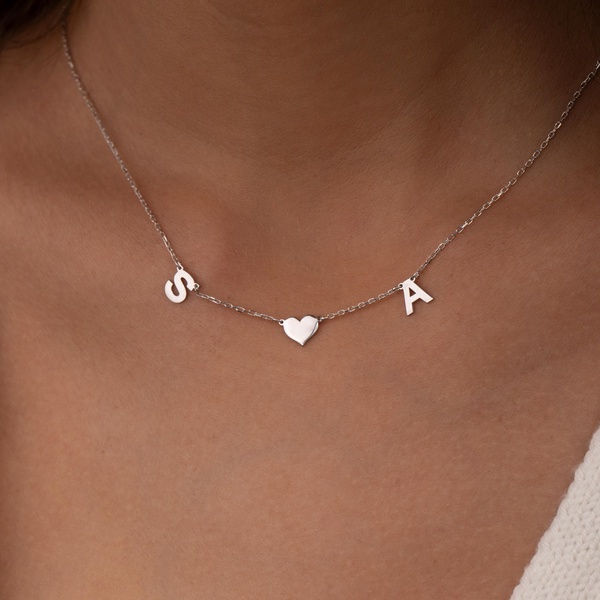 A refined, close-up shot of a silver initial necklace with a central heart charm worn by a person, highlighting product elegance and personal significance.