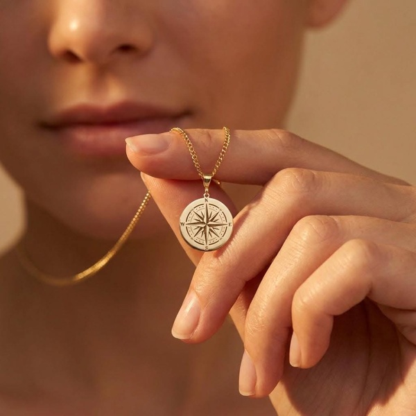 A close-up, high-quality image of a person holding a delicate gold compass pendant, emphasizing the jewelry's intricate craftsmanship and minimalist lifestyle appeal.