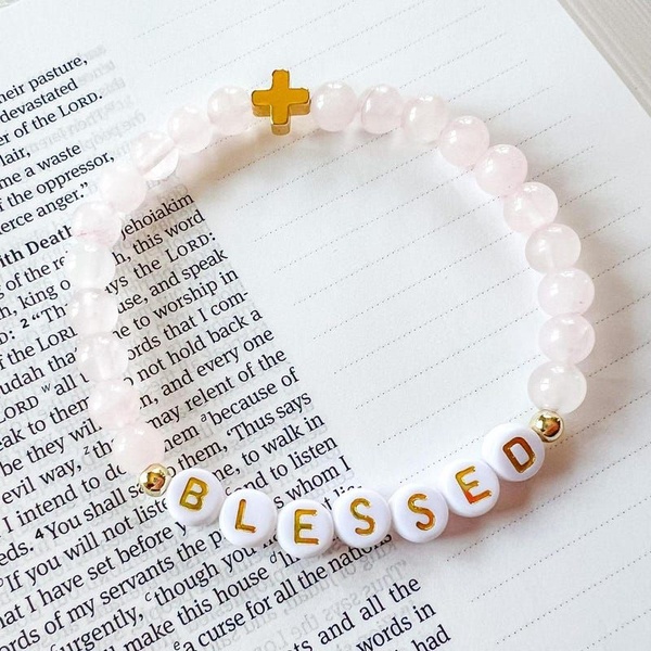 A close-up, high-angle product photograph of a beaded bracelet with the word BLESSED spelled out on letter beads, resting on top of an open book.