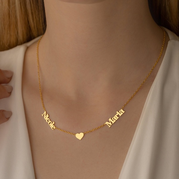 A close-up, high-quality photograph of a gold name necklace featuring two names and a heart charm, worn by a woman.