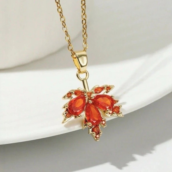 A close-up photograph of a gold maple leaf pendant with vibrant orange gemstone details, displayed elegantly on a white curved surface.