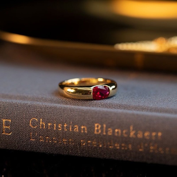 A close-up photograph of a simple gold band set with a single oval ruby, resting on the spine of a book, emphasizing texture and light.
