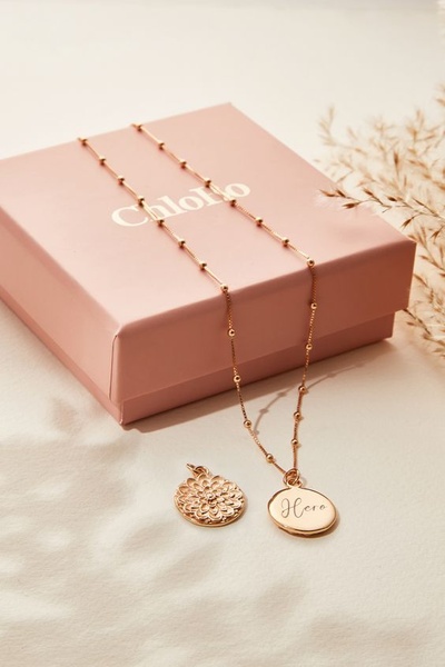 A refined, professional hero shot of a gold necklace with two circular pendants placed elegantly next to its branded gift box.