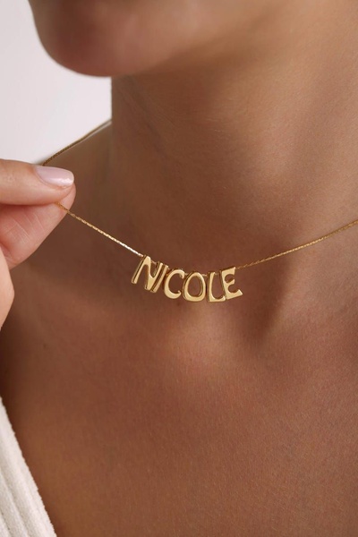 A close-up, high-quality product shot of a gold name necklace worn by a model, emphasizing the clean lines and personalized nature of the jewelry.