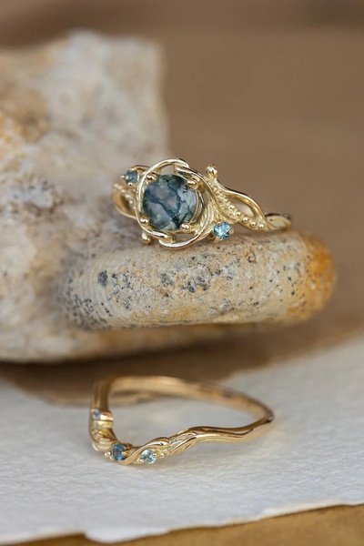 A high-end, close-up product photograph of a gold ring featuring a moss agate center stone and small side accents, resting on a textured stone surface.