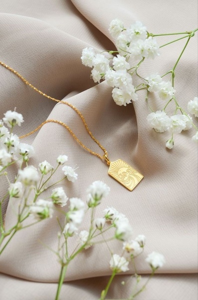 A high-end, minimalist product shot featuring a gold rectangle pendant necklace draped over soft draped fabric and surrounded by baby's breath flowers.