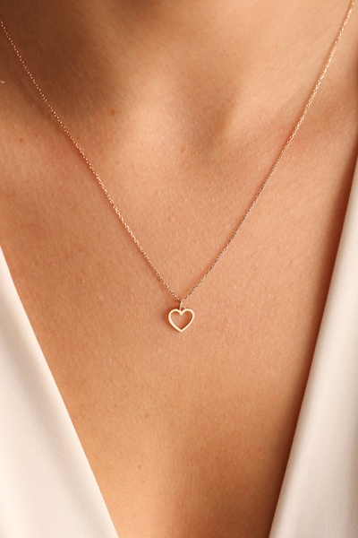 A close-up photograph of a model wearing a delicate gold heart necklace, focusing on product texture and skin tone.
