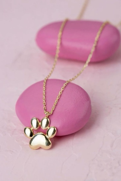 A high-quality studio close-up of a gold paw print pendant necklace centered on soft, pink-toned circular stones.