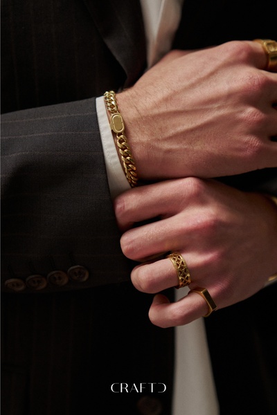 A close-up, high-fashion shot of a person wearing a gold chain bracelet and two gold rings while dressed in a dark suit.