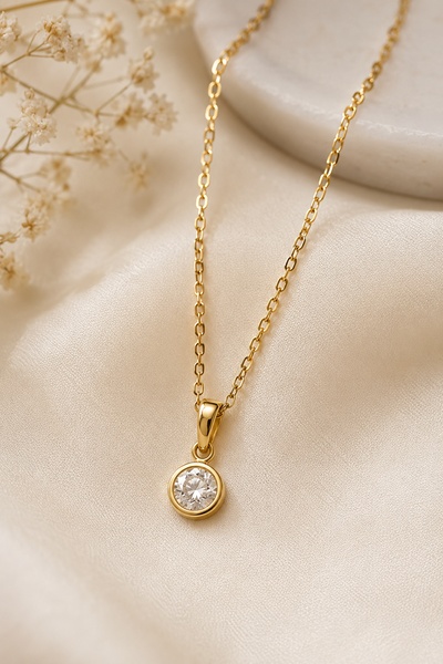 A refined, close-up hero shot of a delicate gold chain necklace with a single round diamond pendant on a soft, textured silk background.