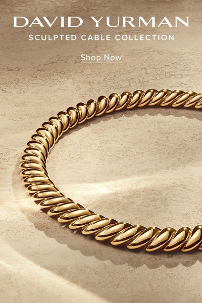 A close-up, high-end product shot of a gold sculpted cable necklace against a neutral, textured stone background.