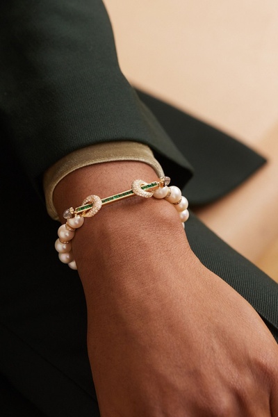 A close-up, high-end product photography shot of a pearl and emerald-accented gold bracelet worn on a wrist against a dark jacket.