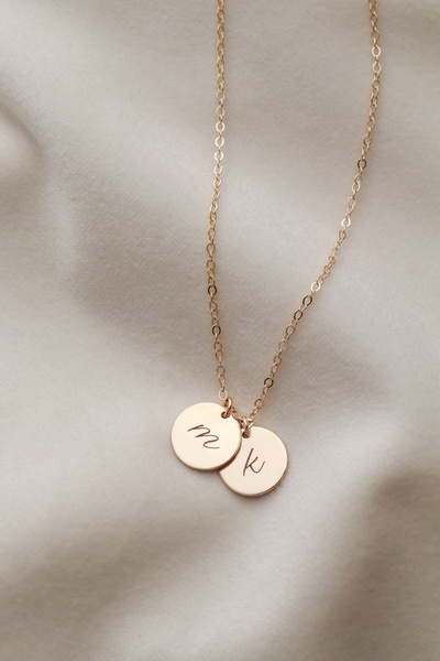 A close-up, soft-focus shot of a delicate gold chain necklace with two circular initial pendants resting on a textured cream fabric backdrop.