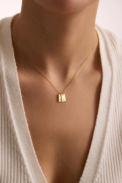 A close-up, minimalist shot of a personalized gold necklace on a model, focusing on the refined texture and custom initial tags.