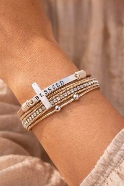 A close-up lifestyle shot of a layered bracelet featuring a cross charm with the word Blessed, set against a warm, soft-focus background.