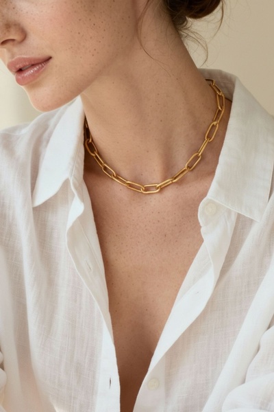 A close-up shot of a model wearing a gold chain necklace with a white collared shirt, emphasizing a clean and high-end aesthetic.