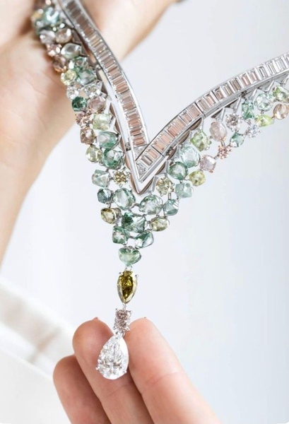 A luxury necklace featuring multicolored gemstones and diamonds is held by a model's hand against a soft-focus background.