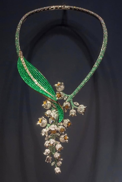 A high-end, artistic close-up of a necklace featuring emerald green and diamond-encrusted lily of the valley flowers against a dark, minimalist backdrop.