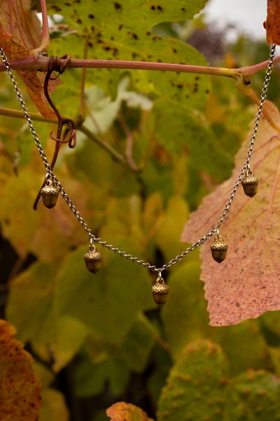This creative features an acorn necklace hanging against a natural autumn backdrop to emphasize seasonal themes and elegant product detail.