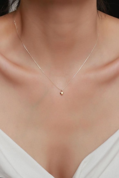 A close-up, high-detail photograph of a delicate gold heart necklace worn by a model against a clean background.