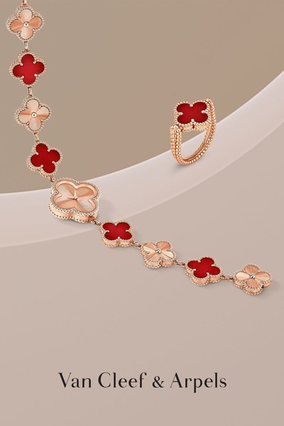 This creative displays a gold necklace and ring featuring a red clover motif, set against a soft, neutral studio backdrop.