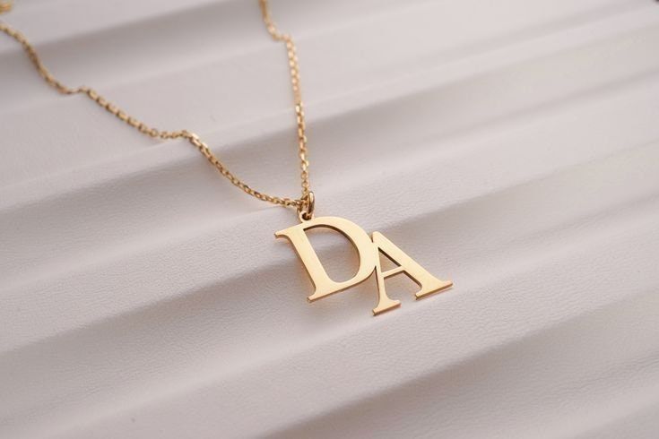 A high-quality studio shot of a gold letter necklace resting on a neutral, textured background.