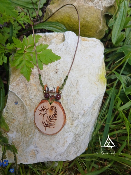 A close-up aesthetic shot of a wooden fern-burned pendant resting on a textured white stone in a natural garden setting.