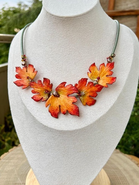 A high-contrast studio shot of a delicate maple leaf necklace displayed on a neutral bust to emphasize the vibrant orange and red tones.