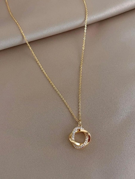 A high-quality, close-up product shot of a gold necklace featuring a circular pendant with inlaid crystals, set against a smooth, neutral fabric background.