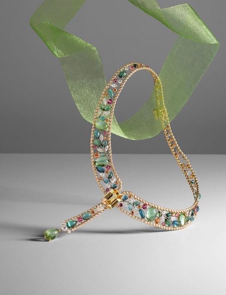 A high-end studio shot of a delicate multi-colored gemstone necklace draped with a green ribbon against a grey background.