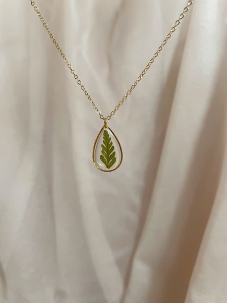 A close-up, top-down shot of a gold tear-drop pendant featuring a preserved fern leaf suspended on a simple gold chain against a soft fabric background.