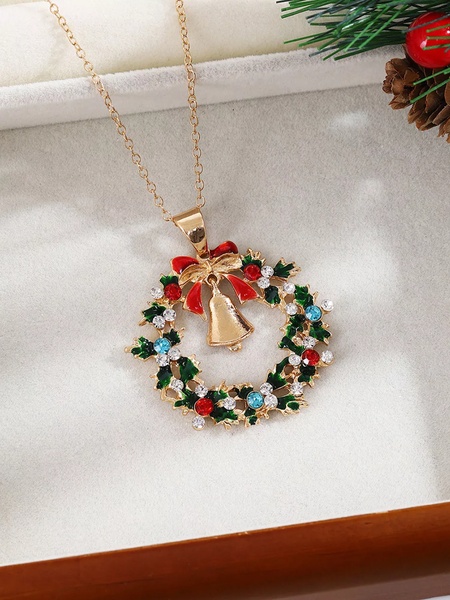 A close-up studio shot of a gold Christmas wreath pendant featuring red, green, and clear stones, resting on a clean white velvet surface.