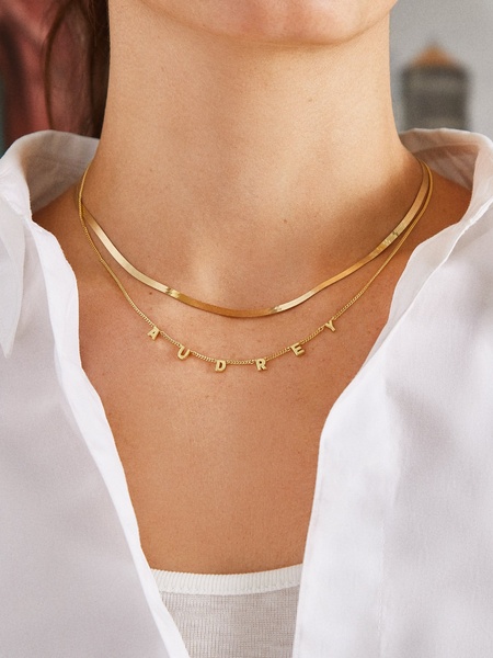 A close-up photograph showcasing a layered gold necklace, with the bottom chain featuring the name Audrey in elegant lettering.