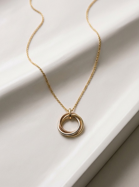 A high-end, clean product shot of a triple-linked ring necklace resting on a soft, draped fabric background.