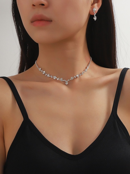 A close-up photograph of a woman wearing a delicate silver-toned crystal necklace and matching earring, showcasing the product in a clean, professional studio environment.