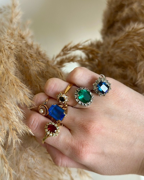 A close-up hand shot displaying five unique fashion rings with different gemstone colors, set against a natural, neutral background.