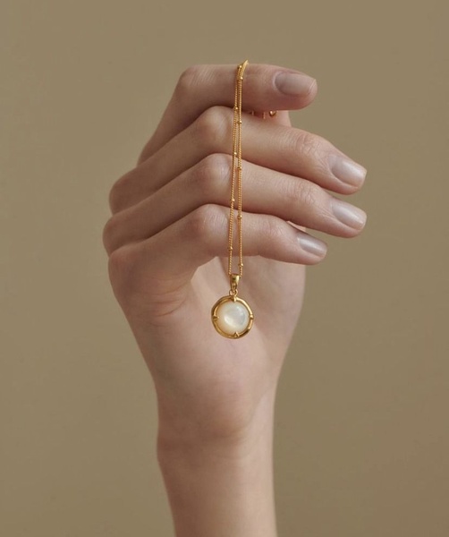 A close-up, high-end studio shot of a hand holding a delicate gold necklace with a pearl pendant, featuring a neutral aesthetic.