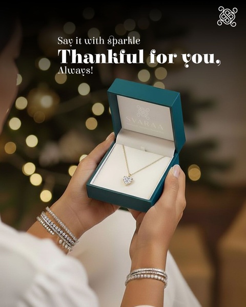 A close-up, first-person perspective showing a woman holding an open box featuring a delicate heart-shaped necklace with a warm, festive bokeh background.