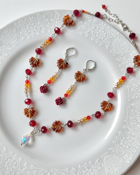 A high-angle flat lay photograph of a leaf-themed necklace and matching earrings featuring red, orange, and amber crystals on a decorative white plate.