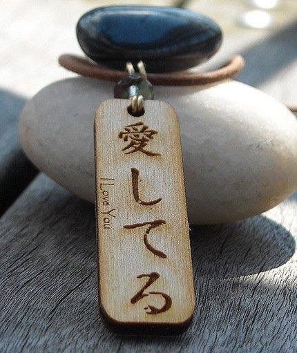 A close-up, high-contrast shot of a wooden pendant inscribed with 'I Love You' in Japanese, resting on smooth beach stones to convey intimacy and organic beauty.