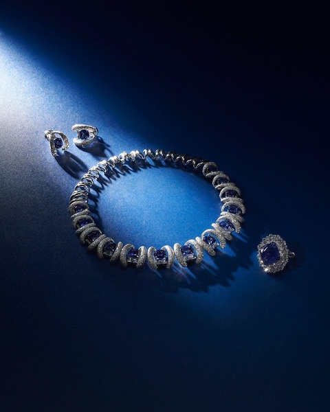 This creative showcases a complete sapphire jewelry set including a necklace, earrings, and ring, arranged elegantly against a dark, moody blue background with dramatic lighting.