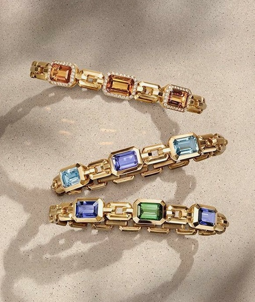 A sophisticated product hero shot featuring three distinct gold-linked bracelets with emerald, sapphire, and citrine gemstones laid out on a textured sand surface.