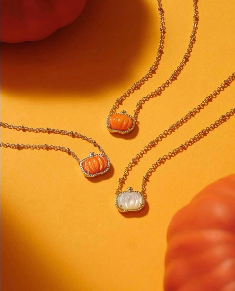 A close-up studio shot featuring three pumpkin-shaped pendant necklaces arranged on a vibrant orange background with pumpkins nearby.