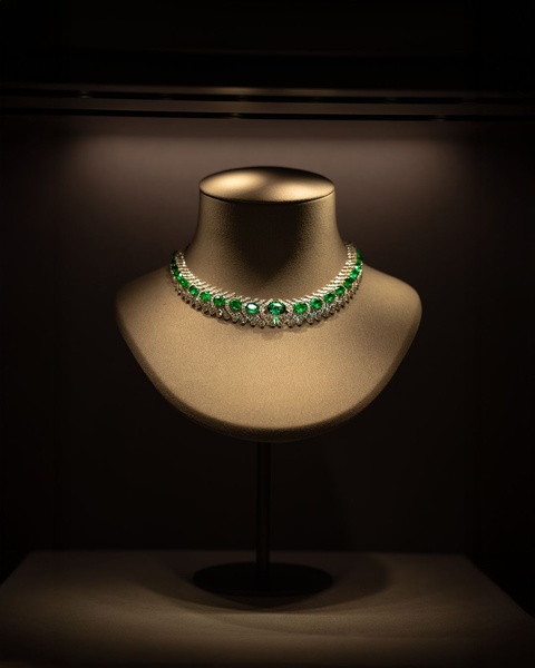 A luxury emerald and diamond necklace is showcased on a neutral-toned mannequin under dramatic, focused spotlighting to highlight its craftsmanship.