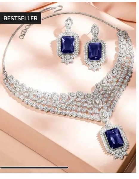 A high-end, clean product hero shot of a sapphire and diamond necklace and earring set displayed against a soft, satin-textured background.